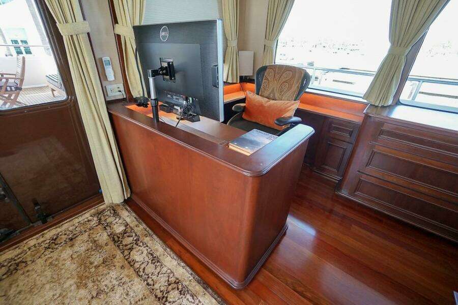 Luxurious yacht interior with wooden desk, monitor, and elegant chair, featuring large windows and curtains.